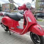 Vespa GT Super 125 cc, 2023 côté droit sur le chevalet central. Moto rouge avec les chromes et la selle noire. Vespa d’occasion Gianni Moto