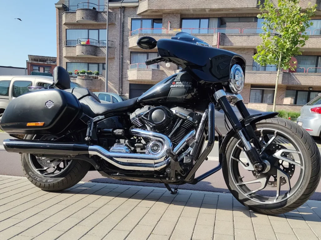 Harley-Davidson Sport Glide noire d'occasion vue de profil droit, avec moteur Milwaukee-Eight chromé, sacoche latérale rigide, échappement noir et carénage Batwing, garée sur un trottoir en Belgique.