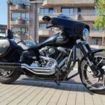 Harley-Davidson Sport Glide noire d'occasion vue de profil droit, avec moteur Milwaukee-Eight chromé, sacoche latérale rigide, échappement noir et carénage Batwing, garée sur un trottoir en Belgique.