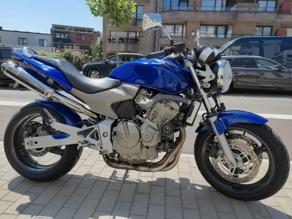 Photographie de profil latéral droit d'une moto naked Honda Hornet CB600F bleu vif avec cadre gris et moteur argenté, garée à l'extérieur sur un trottoir pavé par une journée ensoleillée. L'échappement sport Lazer est visible.