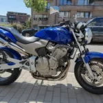 Photographie de profil latéral droit d'une moto naked Honda Hornet CB600F bleu vif avec cadre gris et moteur argenté, garée à l'extérieur sur un trottoir pavé par une journée ensoleillée. L'échappement sport Lazer est visible.