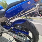 Vue rapprochée du pneu arrière, du bras oscillant gris avec autocollant Lazer, du garde-boue bleu brillant et du silencieux d'échappement en chrome brillant d'une moto Honda Hornet garée sur un trottoir pavé.