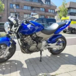 Photographie latérale complète du côté gauche d'une moto Honda Hornet bleue, posée sur sa béquille latérale sur un trottoir pavé. La moto est entièrement visible avec ses jantes noires, son moteur argenté, le silencieux d'échappement sport chromé et les bâtiments urbains à l'arrière-plan. Un fourgon de transport de motos est également visible derrière.
