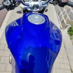 Vue plongeante directe sur le réservoir de carburant d'un bleu métallisé brillant avec les logos Honda argentés, le bouchon de remplissage chromé, le guidon chromé avec ses commandes et leviers noirs, et le tableau de bord à double compteur analogique. L'image est prise sous une lumière naturelle vive.