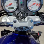 Vue plongeante du poste de pilotage d'une moto Honda Hornet, montrant le tableau de bord à double compteur analogique (indicateur de vitesse à gauche, compte-tours à droite) avec fonds blancs, ainsi que la clé de contact insérée et le té de fourche supérieur en aluminium poli. L'affichage numérique indique un kilométrage de 17893.