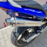 Vue d'angle arrière trois-quarts droit d'une moto Honda Hornet bleue, mettant en évidence le silencieux d'échappement sport Lazer en chrome brillant. On aperçoit également le pneu arrière, le bras oscillant gris mat et le feu arrière rouge intégré dans le carénage bleu. L'autocollant CB600F est visible sur le carénage latéral.