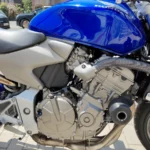 Vue rapprochée détaillée du côté droit du moteur quatre cylindres argenté d'une moto Honda Hornet, montrant les carters de l'embrayage et de l'alternateur, une partie du cadre gris mat et le réservoir de carburant bleu brillant au-dessus. Un tampon de protection moteur noir est visible sur le côté droit.