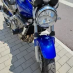 Vue rapprochée d'angle trois-quarts avant de la moto Honda Hornet bleue, mettant en évidence le phare rond chromé, le garde-boue avant bleu brillant, la fourche télescopique avec le disque de frein, une partie du radiateur et du moteur quatre cylindres argenté avec les collecteurs d'échappement. Les tampons de protection moteur noirs sont visibles de ce côté également.